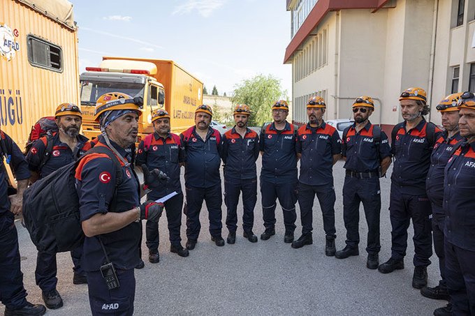 First image shows a group of about ten men wearing orange hard hats, dark blue uniforms with orange accents and Turkish flags on shoulders, standing in a circle on a paved area near yellow shipping containers, orange emergency trucks, and a red building under a clear sky with some trees. Second image depicts a similar group of men in matching uniforms and hard hats gathered around an orange AFAD emergency truck with Turkish flags, some shaking hands or interacting, positioned on a paved surface next to a red building and scaffolding under a blue sky.