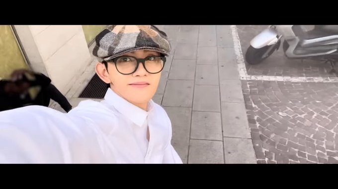 A young man with short black hair smiles at the camera in a selfie, wearing a white shirt, black checkered cap tilted back, and black glasses, standing on a gray tiled outdoor path next to a silver scooter and a black backpack, with beige building walls and green drainage in the background.