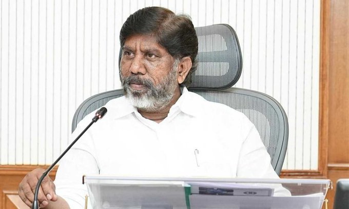 An elderly man with a white beard and hair, wearing a white shirt, sits in a cushioned office chair behind a desk with papers and a microphone in hand, speaking into another microphone on a stand, in a room with wooden paneling and vertical blinds.