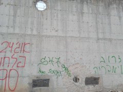 First image shows a row of charred concrete pillars supporting a damaged roof in a debris-filled area with ash and rubble on the ground. Second image depicts a beige concrete building with extensive fire damage including blackened walls a large hole in the upper structure and scattered sand piles nearby. Third image features a concrete wall covered in Hebrew graffiti including numbers 24 and 7 green text and red markings near small window openings. Fourth image displays multiple intact and damaged concrete pillars amid ground littered with plastic bottles bags cardboard boxes and other trash in a dusty environment.