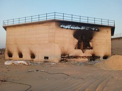 First image shows a row of charred concrete pillars supporting a damaged roof in a debris-filled area with ash and rubble on the ground. Second image depicts a beige concrete building with extensive fire damage including blackened walls a large hole in the upper structure and scattered sand piles nearby. Third image features a concrete wall covered in Hebrew graffiti including numbers 24 and 7 green text and red markings near small window openings. Fourth image displays multiple intact and damaged concrete pillars amid ground littered with plastic bottles bags cardboard boxes and other trash in a dusty environment.