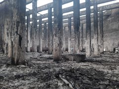 First image shows a row of charred concrete pillars supporting a damaged roof in a debris-filled area with ash and rubble on the ground. Second image depicts a beige concrete building with extensive fire damage including blackened walls a large hole in the upper structure and scattered sand piles nearby. Third image features a concrete wall covered in Hebrew graffiti including numbers 24 and 7 green text and red markings near small window openings. Fourth image displays multiple intact and damaged concrete pillars amid ground littered with plastic bottles bags cardboard boxes and other trash in a dusty environment.
