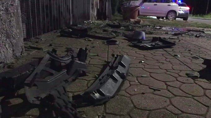 Nighttime scene outside a fenced area with scattered dark debris including metal frames and panels from a destroyed vehicle on a paved ground near potted plants. A white police van with activated blue and red emergency lights is parked nearby on the road. Wet pavement reflects the lights suggesting recent rain or water.