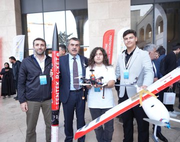 First image shows a large group of formally dressed men and women standing on a blue-lit stage holding awards during a ceremony with a banner reading Imam Hatip Basari Ornekleri Sergisi ve Odul Toreni and screens displaying participants. Second image captures several people including young individuals posing outdoors with a large red rocket model labeled ROCKET, a white drone, and flags in a courtyard setting. Third image features women in headscarves and men around a white vintage car decorated with intricate patterns and a plaque for Kizilay Imam Hatip Ortaokulu Din Geneli 1875 on a patterned floor. Fourth image depicts a man in suit interacting with two children one boy and one girl in headscarf seated in a blue racing simulator cockpit inside a metallic enclosure with screens and sponsor logos like Spar.