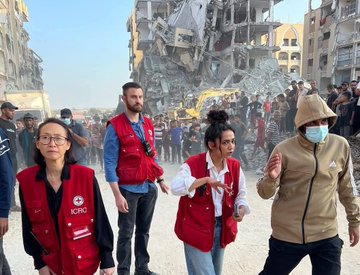 First image displays a large dirt pit filled with debris from destroyed buildings surrounded by damaged structures and scattered rubble with a yellow bulldozer positioned at the edge actively working in the excavation site amid a barren dusty landscape. Second image shows multiple International Committee of the Red Cross workers wearing red vests with ICRC logos standing in a street lined with heavily damaged multi-story buildings collapsed into piles of concrete and rebar with crowds of people including men in hoodies and women in casual clothing gathered nearby observing the scene under a clear sky.