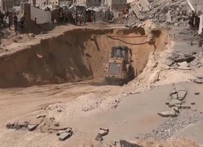 First image displays a large dirt pit filled with debris from destroyed buildings surrounded by damaged structures and scattered rubble with a yellow bulldozer positioned at the edge actively working in the excavation site amid a barren dusty landscape. Second image shows multiple International Committee of the Red Cross workers wearing red vests with ICRC logos standing in a street lined with heavily damaged multi-story buildings collapsed into piles of concrete and rebar with crowds of people including men in hoodies and women in casual clothing gathered nearby observing the scene under a clear sky.