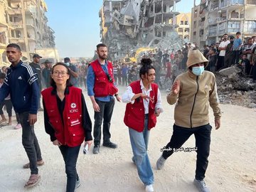 First image shows diverse group of aid workers including women and men in red vests with IRC patches walking through rubble-strewn street amid collapsed buildings and debris under clear sky with onlookers in background. Second image depicts yellow bulldozer excavating large pile of concrete rubble next to damaged structures with several people in red vests standing on elevated debris observing the clearing operation.