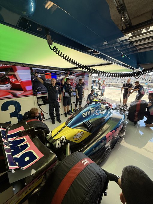 Pit crew members in blue uniforms stand around the teal and blue Alpine F1 car numbered 3 on the pit lane, with green overhead lights on, monitors displaying data, cables and equipment nearby, team gesturing actively in the garage area.