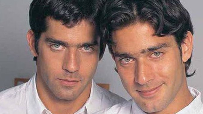 Two men with dark curly hair and green eyes stand side by side wearing white shirts, one with a neutral expression and the other smiling slightly, posed against a plain background.