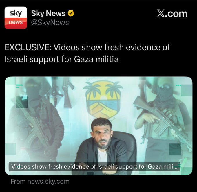 Blurred image shows three masked figures in dark clothing holding rifles standing on either side of a seated man with a beard wearing a light shirt and dark jacket arms crossed in front of a yellow and blue flag with palm trees and a blurred background suggesting a room setting.