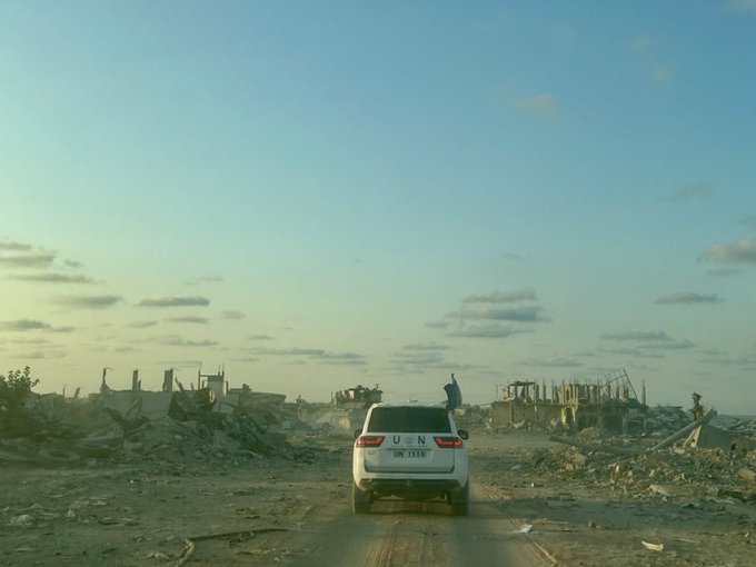 A white SUV with UN markings on the doors and spare tire on the rear drives away on a dusty dirt road flanked by piles of rubble and collapsed concrete structures from destroyed buildings under a sky with scattered clouds during golden hour lighting.