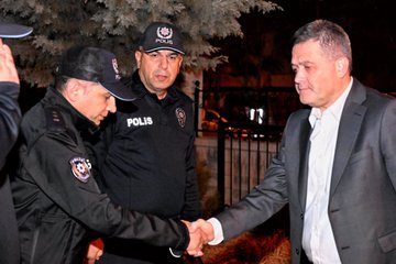 First image shows a man in a gray suit with white shirt standing and talking to a police officer in black tactical vest holding a rifle at night outdoors near a gate and vehicles with orange lighting. Second image depicts three police officers in black uniforms with caps shaking hands with a man in a gray suit under a tree at night outdoors. Third image displays a man in a dark suit sitting at a wooden desk signing documents in an office with two standing police officers in black uniforms nearby a large screen on the wall showing a portrait of Mustafa Kemal Atatürk and Turkish flag emblem. Fourth image illustrates four men including police officers in blue uniforms seated around a wooden table in an office discussing with documents and a bottle on the table near a screen displaying Atatürk portrait and window blinds.
