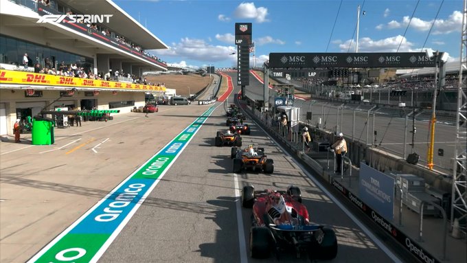 Formula 1 cars in various colors lined up on the starting grid at a racetrack with green and white striped curbs, surrounded by barriers and spectator areas. Grandstands filled with people overlook the scene under a partly cloudy sky. Prominent banners display MSC and SPRINT logos, with a large digital screen tower nearby. The track features asphalt surface with white lines and green astroturf patches adjacent to the barriers.
