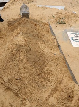 First image displays a sandy cemetery area with a small gray headstone partially buried in disturbed earth, surrounded by sand piles and sparse green plants, with white grave markers bearing Arabic script visible nearby. The ground appears bulldozed and uneven, indicating recent disruption. Second image shows a young infant boy with dark hair and wide eyes, smiling while sitting on a wooden surface, wearing a white short-sleeved onesie, with his hands clasped and a colorful toy nearby.