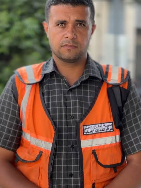 A man with short dark hair and a serious expression stands outdoors wearing an orange high-visibility vest over a gray checkered shirt with a badge on the chest displaying Arabic text. He has a radio clipped to his vest and stands against a background of greenery and buildings.