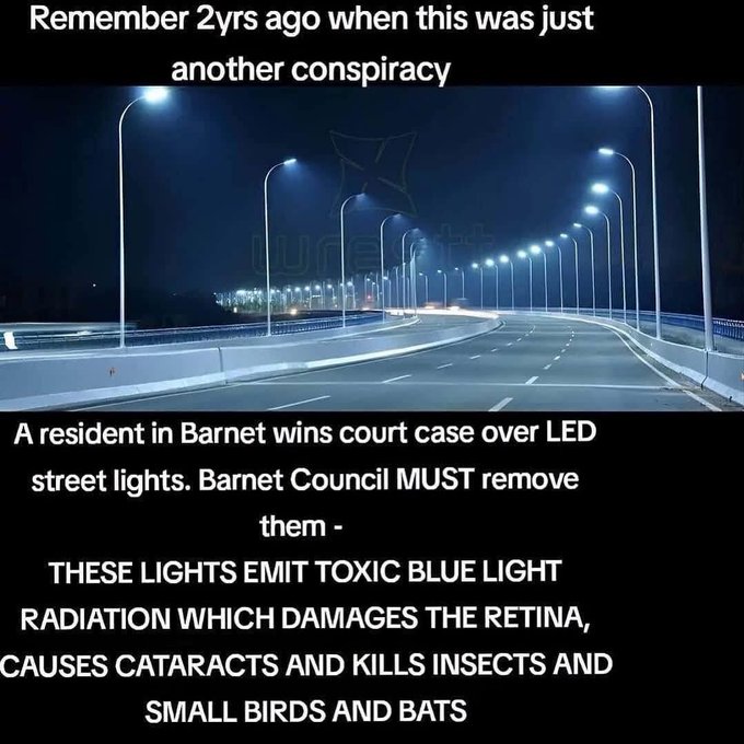 Dark nighttime scene of a straight road flanked by tall metal street lamps emitting bright white light from LED bulbs spaced evenly along the curbs with a bridge structure visible ahead and minimal traffic under overcast sky.