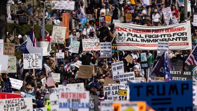 Large crowd of people gathered at an outdoor protest rally holding numerous signs with messages such as Abolish ICE Stop Deportations Newsom for President Full Rights for All Immigrants Free Palestine and Justice and Freedom Party banners visible along with American flags and a Trump sign some participants wearing hats and casual clothing trees and urban setting in the background