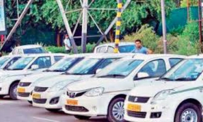 Multiple white sedans resembling taxis parked in a row along a roadside with yellow license plates visible on some vehicles. A few men stand near the cars one wearing a white shirt and another in a blue outfit. Green trees and foliage surround the area with metal poles and a fence in the background under clear daylight.