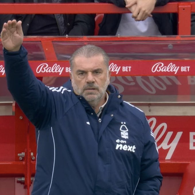 A bald man with a beard wearing a dark blue Nottingham Forest jacket with white stripes and sponsor logos stands in a stadium stand raising his right hand palm out gesturing toward the field with a serious expression surrounded by blurred spectators in red seating areas and Ballys Bet advertising on barriers.