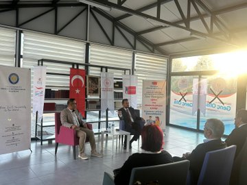 First image shows panel discussion with four people seated on chairs in a modern hall with Turkish flags banners and EU symbols under sunlight through windows. Second image depicts speaker addressing audience of about 20 young people in suits and casual attire seated in rows in a bright room with glass walls and plants. Third image captures group selfie with around 25 diverse young participants smiling in a library-like setting with bookshelves and whiteboards. Fourth image features man in suit receiving framed painting from woman in traditional attire next to chairs and banners in an open indoor space.