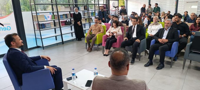 First image shows panel discussion with four people seated on chairs in a modern hall with Turkish flags banners and EU symbols under sunlight through windows. Second image depicts speaker addressing audience of about 20 young people in suits and casual attire seated in rows in a bright room with glass walls and plants. Third image captures group selfie with around 25 diverse young participants smiling in a library-like setting with bookshelves and whiteboards. Fourth image features man in suit receiving framed painting from woman in traditional attire next to chairs and banners in an open indoor space.