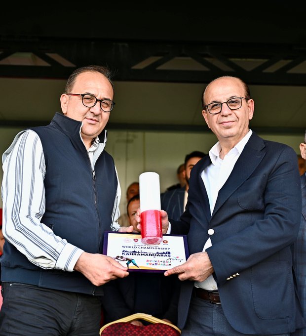 Two men in formal suits stand side by side holding a framed plaque labeled World Karakucak Championship Kahramanmaraş with a red ribbon. One man holds a red-handled white candle. Background shows an outdoor stadium setting with blurred spectators and a red cushioned chair.