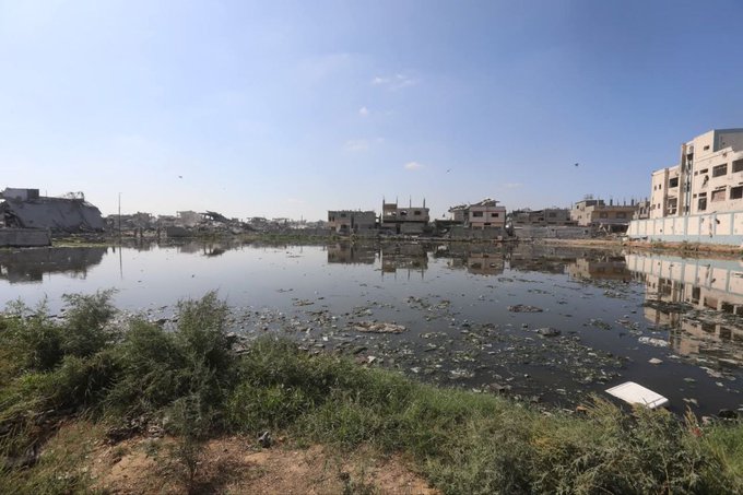 First image shows rusted metal structures, pipes, and hoses connected to a facility amid debris and partial flooding under a clear sky. Second image depicts a white truck with pumping equipment and hoses extending into water near damaged buildings and a lamppost. Third image captures a large body of polluted water surrounded by ruined multi-story buildings and sparse vegetation. Fourth image illustrates a collapsed multi-story building with exposed interiors, debris piles, and surrounding floodwater reflecting the structure.
