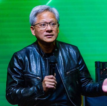 An older Asian man with gray hair and glasses wearing a black textured leather jacket holds a microphone while speaking on stage against a green backdrop with lighting effects and equipment visible nearby. A red flag with five yellow stars arranged in a specific pattern representing the national flag of China.