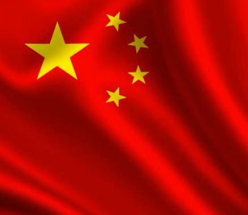 An older Asian man with gray hair and glasses wearing a black textured leather jacket holds a microphone while speaking on stage against a green backdrop with lighting effects and equipment visible nearby. A red flag with five yellow stars arranged in a specific pattern representing the national flag of China.