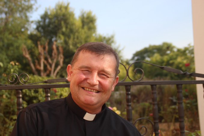 Smiling middle-aged man with short brown hair wearing black clerical shirt and white collar stands outdoors in front of ornate black metal fence with green plants and trees in background under blue sky. Another image shows same man in clerical attire at outdoor protest holding white sign surrounded by crowd with Spanish flags women and men some wearing glasses.
