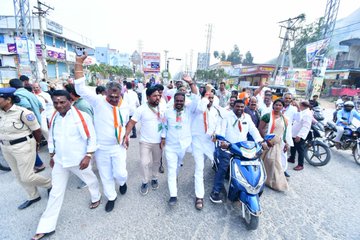 First image displays group of men and boys near school building with Telugu signage on wall surrounded by trees some wearing white shirts and orange scarves others in school uniforms with backpacks standing beside scooter. Second image captures crowd of men in white kurtas and scarves raising arms in gesture near roadside shops and parked scooters with police in uniform present. Third image shows man in white attire with orange scarf speaking into microphone at outdoor event under large tree with seated audience in chairs many wearing similar scarves and traditional clothing.