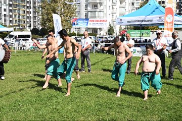 First image shows a group of men in suits standing on a stadium field, several holding and waving a large Turkish flag with red background and white crescent moon and star, others clapping, background includes green barriers and seated audience. Second image depicts a man in dark suit and glasses speaking into a microphone at a white podium decorated with ornate patterns, surrounded by media microphones from various outlets including red ones, backdrop with blue banner reading 20 ULUSLARARASI TURGUT ÖZAL KARAKUÇAK GÜREŞLERİ and image of Turgut Özal, green tent and crowd in background. Third image captures several men and boys in green baggy pants performing traditional wrestling or dance moves on green field, some shirtless with muscular builds, others overweight, near blue tent with YEŞILYEŞIL YEŞILYEŞIL YEŞILYEŞIL text, vehicles and apartments in background. Fourth image is an aerial view of a park area with white domed building, surrounding apartment buildings, event setup with blue tents, flags, crowds of people in white and colored attire, cars parked along roads.