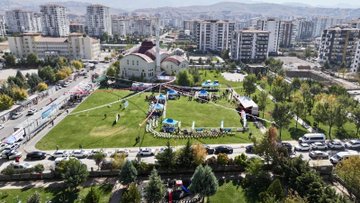 First image shows a group of men in suits standing on a stadium field, several holding and waving a large Turkish flag with red background and white crescent moon and star, others clapping, background includes green barriers and seated audience. Second image depicts a man in dark suit and glasses speaking into a microphone at a white podium decorated with ornate patterns, surrounded by media microphones from various outlets including red ones, backdrop with blue banner reading 20 ULUSLARARASI TURGUT ÖZAL KARAKUÇAK GÜREŞLERİ and image of Turgut Özal, green tent and crowd in background. Third image captures several men and boys in green baggy pants performing traditional wrestling or dance moves on green field, some shirtless with muscular builds, others overweight, near blue tent with YEŞILYEŞIL YEŞILYEŞIL YEŞILYEŞIL text, vehicles and apartments in background. Fourth image is an aerial view of a park area with white domed building, surrounding apartment buildings, event setup with blue tents, flags, crowds of people in white and colored attire, cars parked along roads.