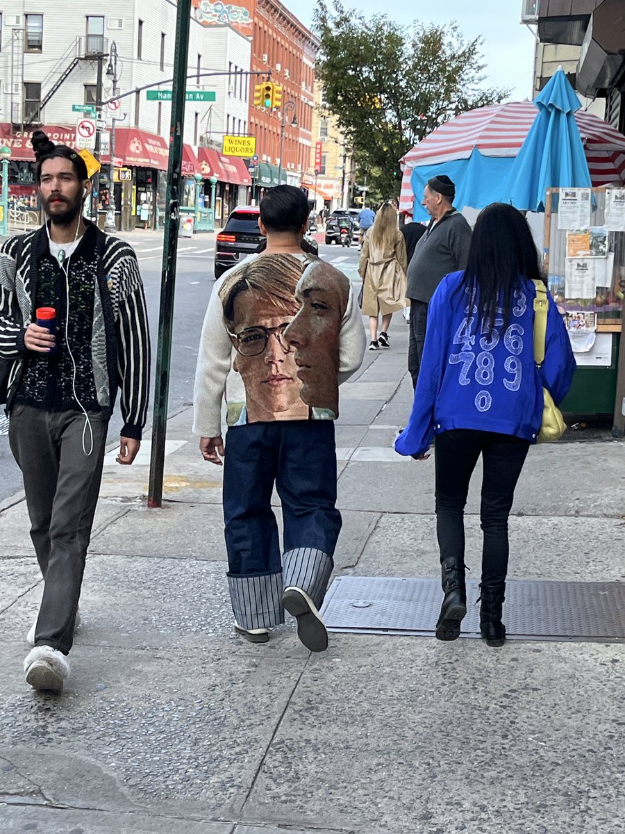 Urban street with brick buildings, traffic lights, and shops in background. Group of people walking: man in black and white striped jacket, red drink in hand, wide-leg denim pants, and sneakers; another man in light sweater with large front print of a mans face wearing glasses, light pants, and shoes; woman in blue numbered 1989 hoodie, yellow bag, black pants, and boots; others in casual wear including beige coat and gray hoodie. Red and white striped umbrella and blue umbrella visible near vendors.