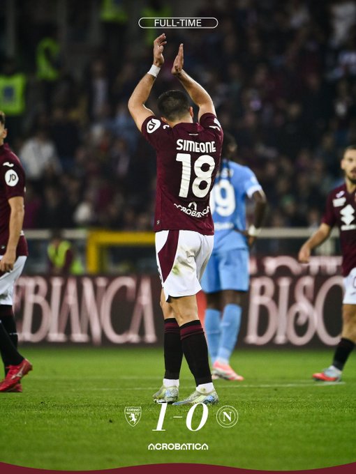 A soccer player in a maroon Torino jersey with number 18 on the back stands on the field raising both arms in celebration with wrists wrapped while facing away from the camera other players in similar maroon kits and blue Napoli kits are nearby in the background stadium seating and sponsor banners like Biancacasa are visible on the pitch edges a full-time graphic overlay shows Torino 1-0 Napoli with team crests.