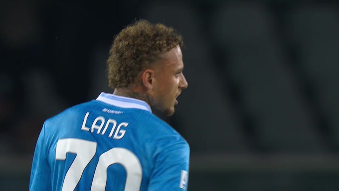Profile view of a male soccer player with curly blond hair wearing a blue Napoli jersey with white accents, number 70, and name LANG on back, standing on a green field with blurred stadium seating in background.