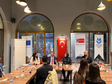 First image shows a group of people including men and women in professional attire seated around a wooden table in a room with arched windows and Turkish and EU flags on walls discussing with water bottles and glasses present. Second image depicts a larger group standing together holding a plaque with Turkish flag and banners in background near tables with drinks. Third image features multiple individuals including suited men women and a child posing in front of a building entrance with green logo and trees on cobblestone path. Fourth image captures people gathered outdoors at night near a banner under trees with some in formal wear engaging in conversation on paved area.