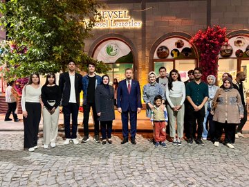 First image shows a group of people including men and women in professional attire seated around a wooden table in a room with arched windows and Turkish and EU flags on walls discussing with water bottles and glasses present. Second image depicts a larger group standing together holding a plaque with Turkish flag and banners in background near tables with drinks. Third image features multiple individuals including suited men women and a child posing in front of a building entrance with green logo and trees on cobblestone path. Fourth image captures people gathered outdoors at night near a banner under trees with some in formal wear engaging in conversation on paved area.
