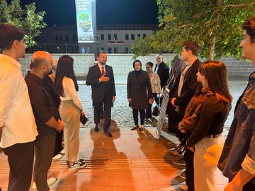 First image shows a group of people including men and women in professional attire seated around a wooden table in a room with arched windows and Turkish and EU flags on walls discussing with water bottles and glasses present. Second image depicts a larger group standing together holding a plaque with Turkish flag and banners in background near tables with drinks. Third image features multiple individuals including suited men women and a child posing in front of a building entrance with green logo and trees on cobblestone path. Fourth image captures people gathered outdoors at night near a banner under trees with some in formal wear engaging in conversation on paved area.