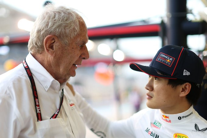 An elderly man with white hair wearing a white shirt lanyard and credentials stands facing a younger Asian man in a red New Era cap white racing suit with sponsor patches including Red Bull Mobil 1 and wings logo they appear to be talking in a garage area with red barriers lighting and blurred background elements