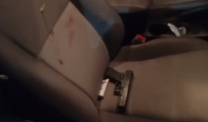 A handgun rests on a beige car seat inside a vehicle with red blood stains visible on the adjacent seat fabric and headrest a red cup sits in the cup holder and a small object is on the floor near the seat base