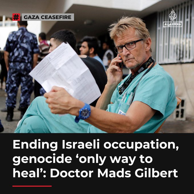 A blonde man wearing glasses and a blue surgical cap sits on a chair holding a phone to his ear while examining papers in his other hand. He is dressed in a blue medical gown and has a blue wristwatch. Several people in military uniforms and civilians stand around in a busy outdoor area with buildings and fences in the background. An Al Jazeera logo appears in the corner.