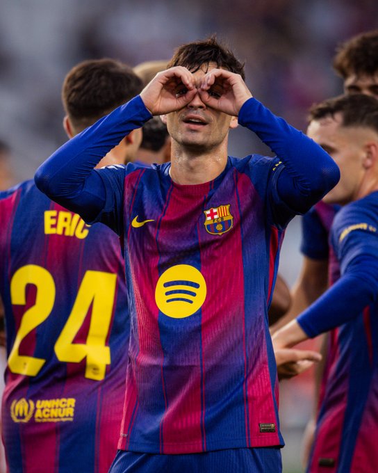 Pedri in FC Barcelona jersey with number 8 and Spotify sponsor, making OK gesture over eyes with hands, surrounded by teammates in matching blue and red uniforms on soccer field in stadium with crowd.