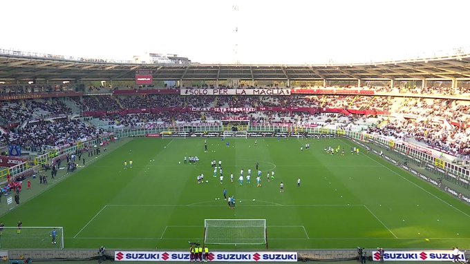 Wide-angle view of a large soccer stadium in Turin with green field marked for play surrounded by multi-tiered stands partially filled with spectators in red and white sections banners reading PER LA MAGLIA visible on upper levels goalposts at ends advertising banners for Suzuki along sidelines.