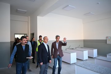 First image shows group of men in professional attire including vests and suits standing and gesturing in spacious indoor area with white walls, ceiling lights, multiple white sinks and counters along walls, and open doorways. Second image depicts group of people in dark clothing gathered on steps in front of large modern building with orange and white facade, green accents, large windows, and paved courtyard. Third image features several men in casual and formal wear walking across paved outdoor area toward multi-story building with orange and green walls, white sections, and surrounding buildings in background. Fourth image is aerial view of single-story school building with red tiled roof, orange and white walls, green sections, entrance stairs, small group of people below, and nearby apartment buildings and road.