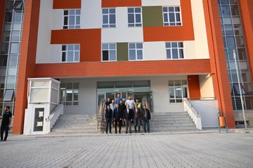 First image shows group of men in professional attire including vests and suits standing and gesturing in spacious indoor area with white walls, ceiling lights, multiple white sinks and counters along walls, and open doorways. Second image depicts group of people in dark clothing gathered on steps in front of large modern building with orange and white facade, green accents, large windows, and paved courtyard. Third image features several men in casual and formal wear walking across paved outdoor area toward multi-story building with orange and green walls, white sections, and surrounding buildings in background. Fourth image is aerial view of single-story school building with red tiled roof, orange and white walls, green sections, entrance stairs, small group of people below, and nearby apartment buildings and road.