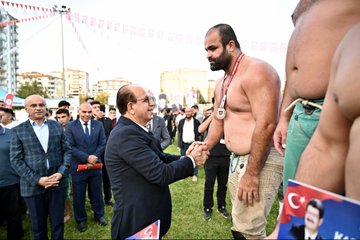 First image shows six men on a stage with Turkish flags and banners for Yeşilyurt and wrestling event three shirtless muscular wrestlers wearing medals and green shorts raising arms in victory two suited older men in center one bald with glasses wearing medal and green pants another in suit all smiling against blue backdrop with sponsor logos. Second image depicts a suited bald man with glasses shaking hands with a bearded shirtless wrestler wearing gold medal and green pants on grassy field surrounded by crowd in formal attire Turkish flags and event banners in background. Third image features group of eight men on grassy area with podium three shirtless wrestlers with medals raising arms two suited men in center including bald glasses wearer others in vests and shirts against backdrop with Turkish flags and event text. Fourth image is aerial view of large grassy park area with multiple white tents colorful flags and banners including large portraits of a man and Turkish flags cars parked around apartment buildings and mountains in distance under sunny sky.