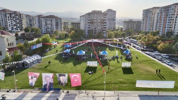 First image shows six men on a stage with Turkish flags and banners for Yeşilyurt and wrestling event three shirtless muscular wrestlers wearing medals and green shorts raising arms in victory two suited older men in center one bald with glasses wearing medal and green pants another in suit all smiling against blue backdrop with sponsor logos. Second image depicts a suited bald man with glasses shaking hands with a bearded shirtless wrestler wearing gold medal and green pants on grassy field surrounded by crowd in formal attire Turkish flags and event banners in background. Third image features group of eight men on grassy area with podium three shirtless wrestlers with medals raising arms two suited men in center including bald glasses wearer others in vests and shirts against backdrop with Turkish flags and event text. Fourth image is aerial view of large grassy park area with multiple white tents colorful flags and banners including large portraits of a man and Turkish flags cars parked around apartment buildings and mountains in distance under sunny sky.