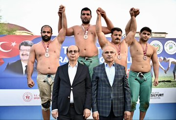 First image shows six men on a stage with Turkish flags and banners for Yeşilyurt and wrestling event three shirtless muscular wrestlers wearing medals and green shorts raising arms in victory two suited older men in center one bald with glasses wearing medal and green pants another in suit all smiling against blue backdrop with sponsor logos. Second image depicts a suited bald man with glasses shaking hands with a bearded shirtless wrestler wearing gold medal and green pants on grassy field surrounded by crowd in formal attire Turkish flags and event banners in background. Third image features group of eight men on grassy area with podium three shirtless wrestlers with medals raising arms two suited men in center including bald glasses wearer others in vests and shirts against backdrop with Turkish flags and event text. Fourth image is aerial view of large grassy park area with multiple white tents colorful flags and banners including large portraits of a man and Turkish flags cars parked around apartment buildings and mountains in distance under sunny sky.