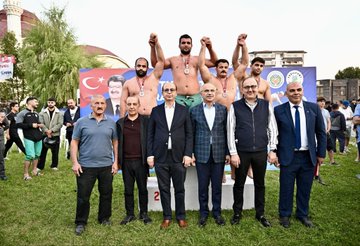 First image shows six men on a stage with Turkish flags and banners for Yeşilyurt and wrestling event three shirtless muscular wrestlers wearing medals and green shorts raising arms in victory two suited older men in center one bald with glasses wearing medal and green pants another in suit all smiling against blue backdrop with sponsor logos. Second image depicts a suited bald man with glasses shaking hands with a bearded shirtless wrestler wearing gold medal and green pants on grassy field surrounded by crowd in formal attire Turkish flags and event banners in background. Third image features group of eight men on grassy area with podium three shirtless wrestlers with medals raising arms two suited men in center including bald glasses wearer others in vests and shirts against backdrop with Turkish flags and event text. Fourth image is aerial view of large grassy park area with multiple white tents colorful flags and banners including large portraits of a man and Turkish flags cars parked around apartment buildings and mountains in distance under sunny sky.